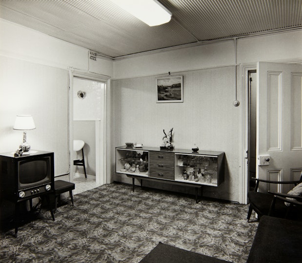 A carpeted, fluorescent-lit living room with a TV, sideboard and armchair.