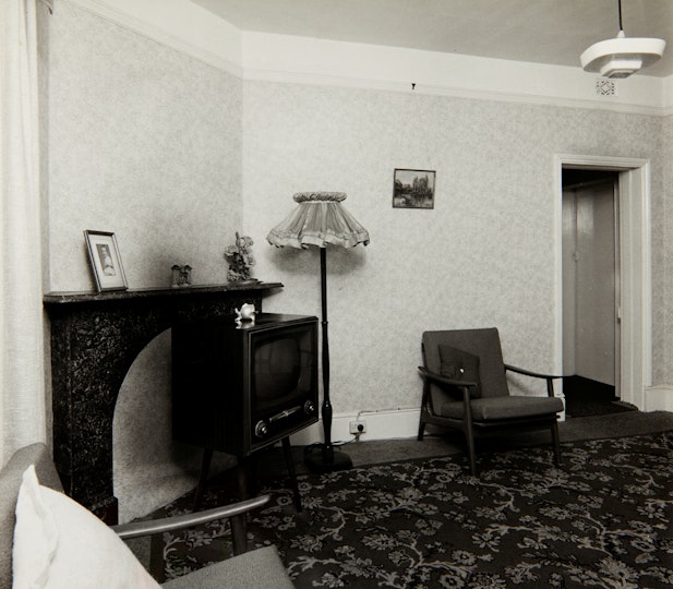 A carpeted living room with an armchair, standing lamp and mantlepiece.