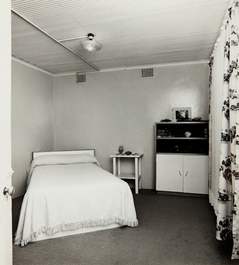 A bedroom with a double bed, bedside table, shelving unit and curtained window.