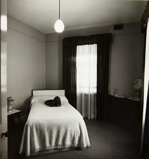 A bedroom with a single bed, bedside table, dresser and two curtained windows.