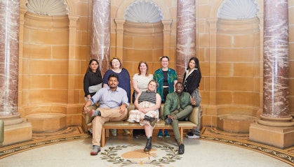 Eight people (three seated, five standing behind them) pose in a room with sandstone wall, marble columns and mosaic floor.