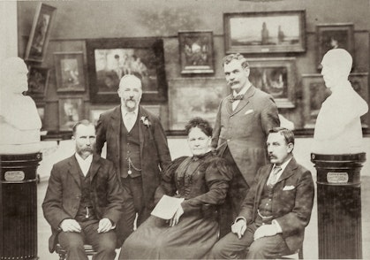 In front of a wall of paintings and between two busts on plinths, a woman sits surrounding by two seated and two standing men.