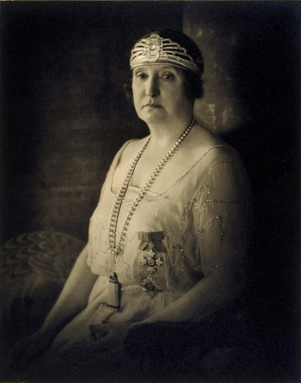 A seated woman dressed in a chiffon dress with sparkly embroidery. She wears a ribboned medal on her chest, long necklaces and a jewelled headpiece around her bobbed hair.
