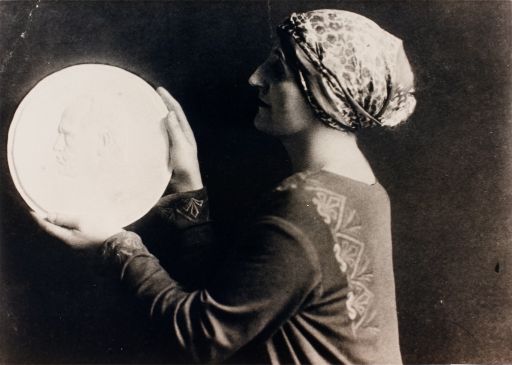 A person with a patterned headwrap holds up a large circular disc depicting a head in relief.