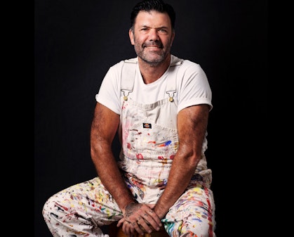 A seated person wearing a white t-shirt and paint-spattered white overalls.