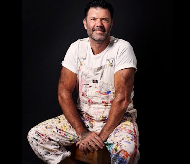 A seated person wearing a white t-shirt and paint-spattered white overalls.