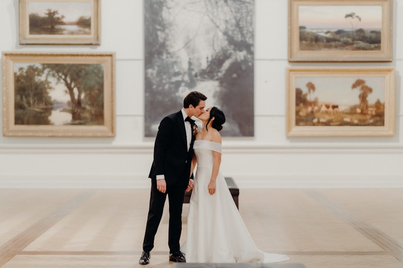 A couple kissing in front of five paintings on a wall in an historic gallery space.