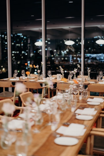 Tables set for dinner in a restaurant with city lights visible through large glass windows.
