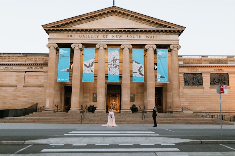 A person in a long white dress and a person in a suit stand in front of a large sandstone building with 'Art Gallery of New South Wales' written above columns and banners at the entrance.