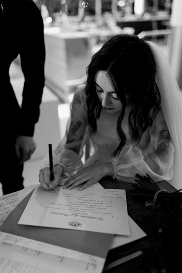 A person with long dark hair and a white veil signs a document on a table.