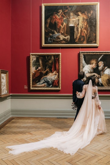 Two people stand with their arms around in other in front of paintings on a wall in an historic gallery space.