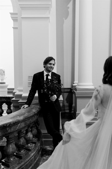 A person in a suit and tie leans against a balustrade. Facing them is a person holding out the skirt of their long dress.