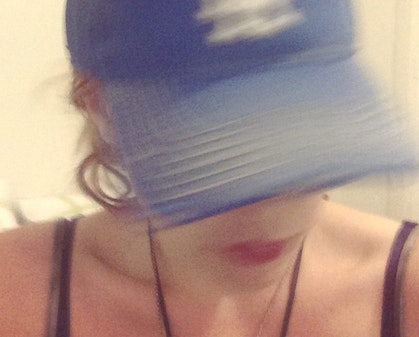 A blurred photo of a person wearing a blue baseball cap, which is obscuring most of their face, except for their mouth.