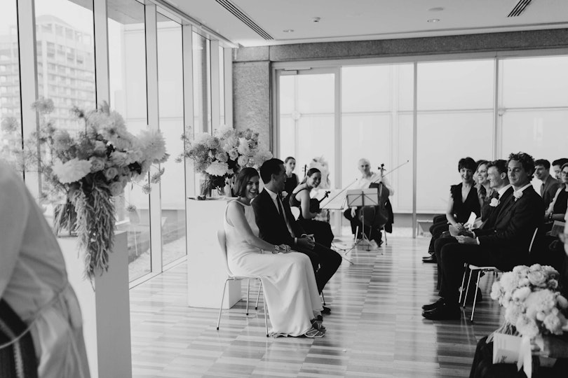 A couple are seated between floral arrangements in a room with floor to ceiling windows in front of seated guests. Three musicans are seated in the corner.