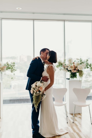Two people embrace in a room in front of large floral arrangements. One person wears a suit. The other wears a white dress and carries a bouquet.