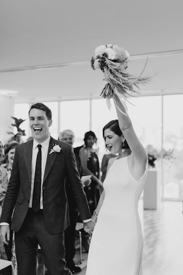 Two people walking in a room, hand in hand. One of them holds up a floral bouquet.