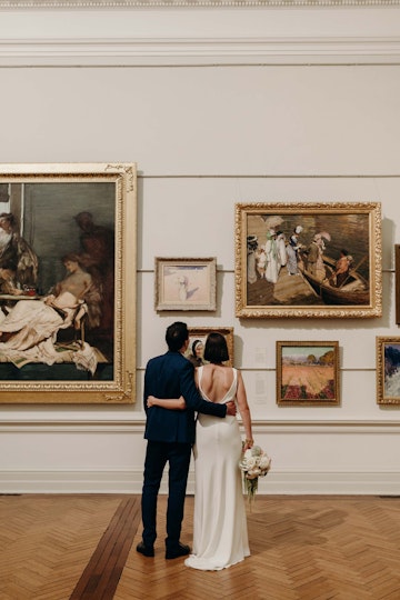 Two people stand with their arms around in other in front of paintings on a wall in an historic gallery space.