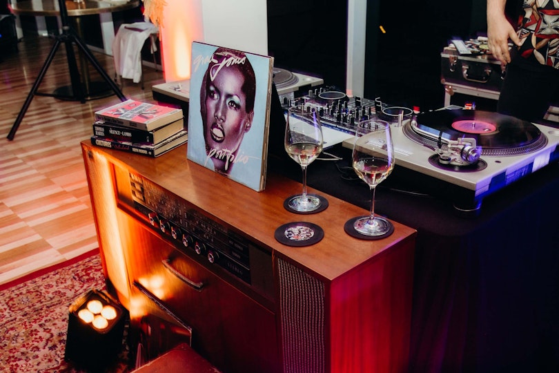 Turntables and other DJ equipment along with a Grace Jones album, a pile of three books and two glasses of wine.