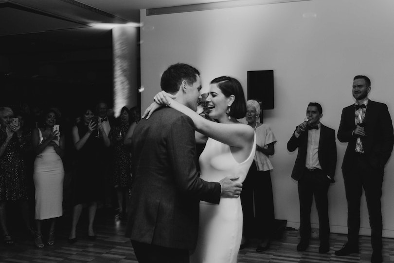 Two people dance with their arms around each others as other people look on.