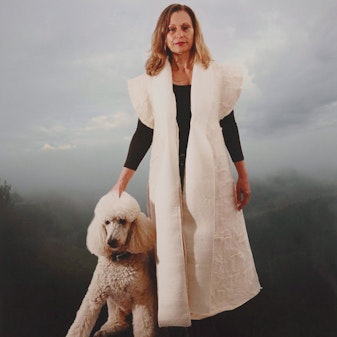 A person in a long short-sleeved white coat over a long-sleeved dark top, with their hand on a white poodle.