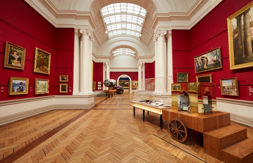 A red-walled gallery hung with paintings with a series of sculptural objects and display cases displayed on the wooden floor