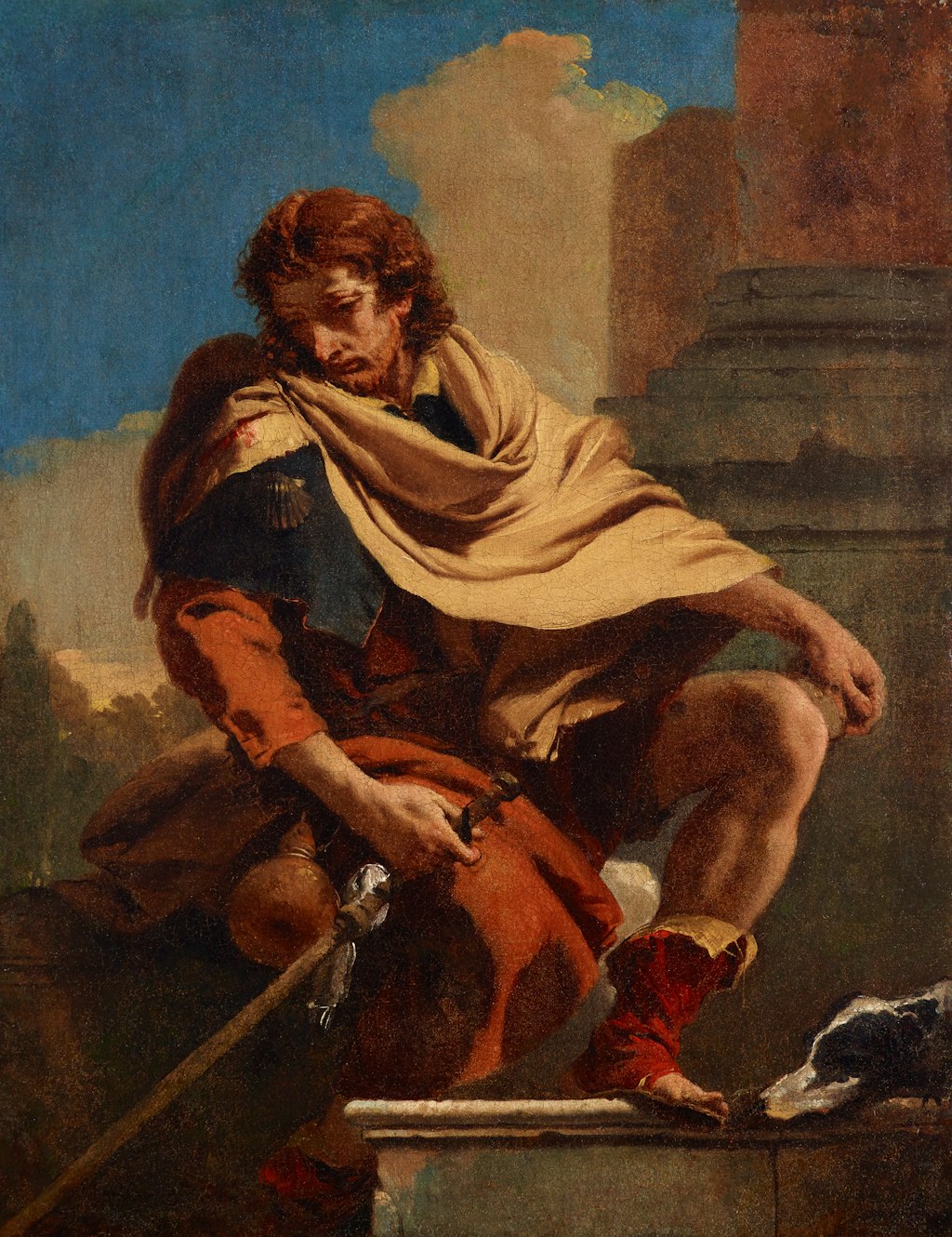 A person with a cloak around their shoulders, holding a staff, sits outside a building, with a dog at their foot.