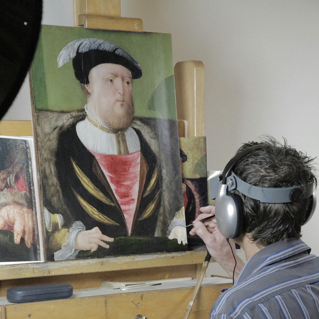 A person with a small tool in their right hand works on a portrait painting on an easel.
