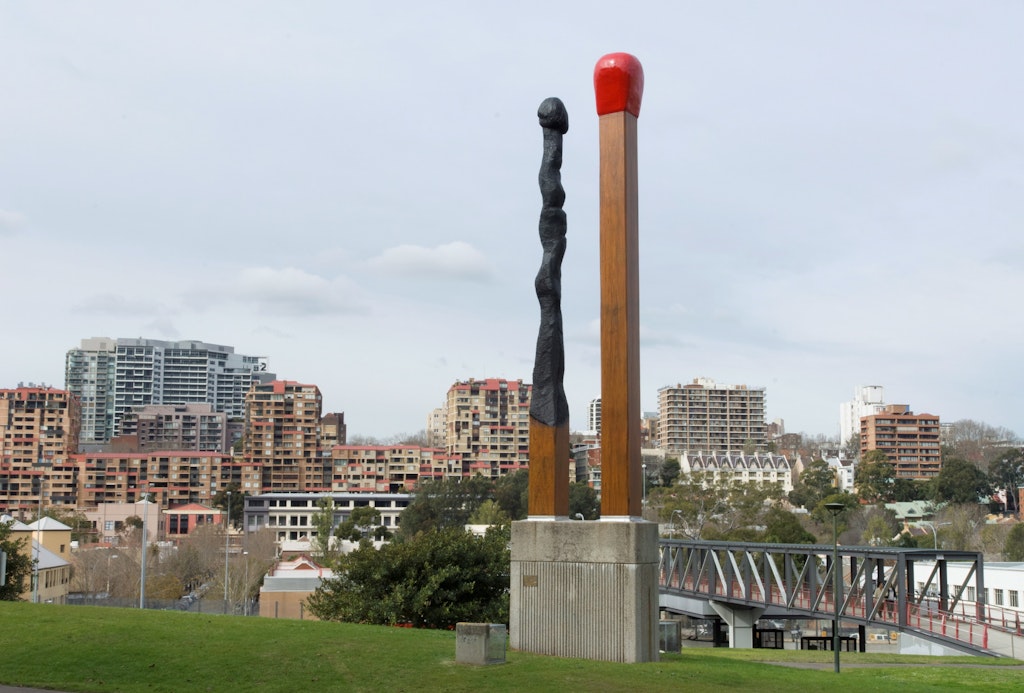 Two giant matchsticks stand on a concret plinth on grass, with buildings in the background. One match is straight and red-tipped. The other appears blackened and burnt.