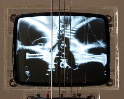 A black-and-white television monitor in translucent casing with four metal strings vertically across its centre.