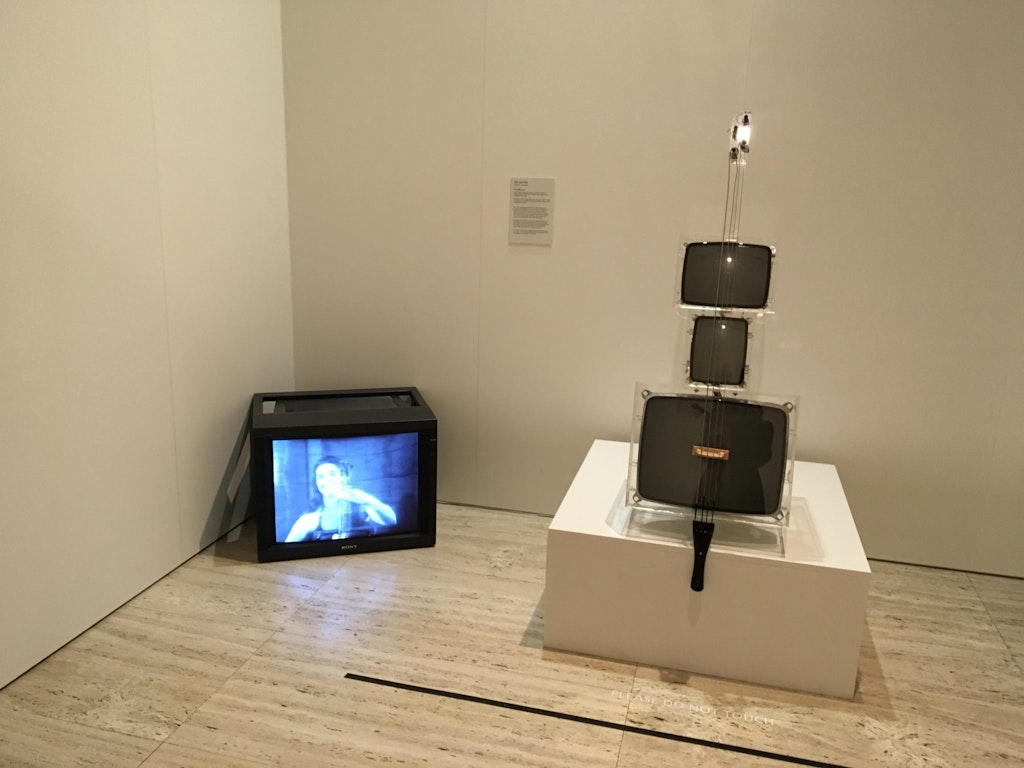 A TV monitor in a black case sits on the floor in the corner. Next to it on a plinth is a cello-like object made up of three monitors.