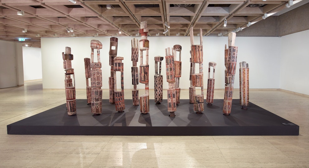 Fifteen carved and painted poles stand on a plinth in a gallery space.