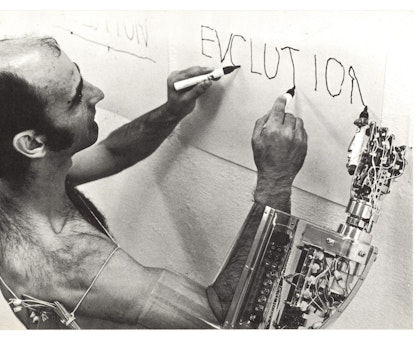 A person wearing a mechanical arm uses their own two hands and the mechanical hand to write 'evolution' in black marker on a sheet of paper on a wall.