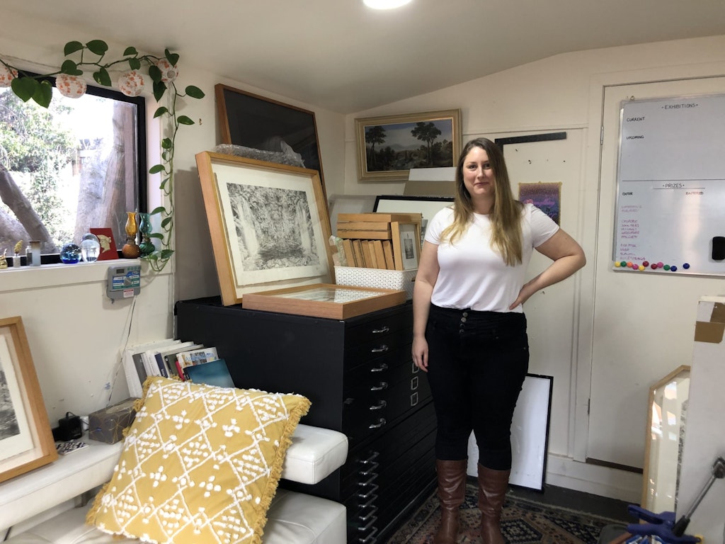 A person stands in a room where framed works of art sit on filing cabinets, the back of a sofa and the floor.