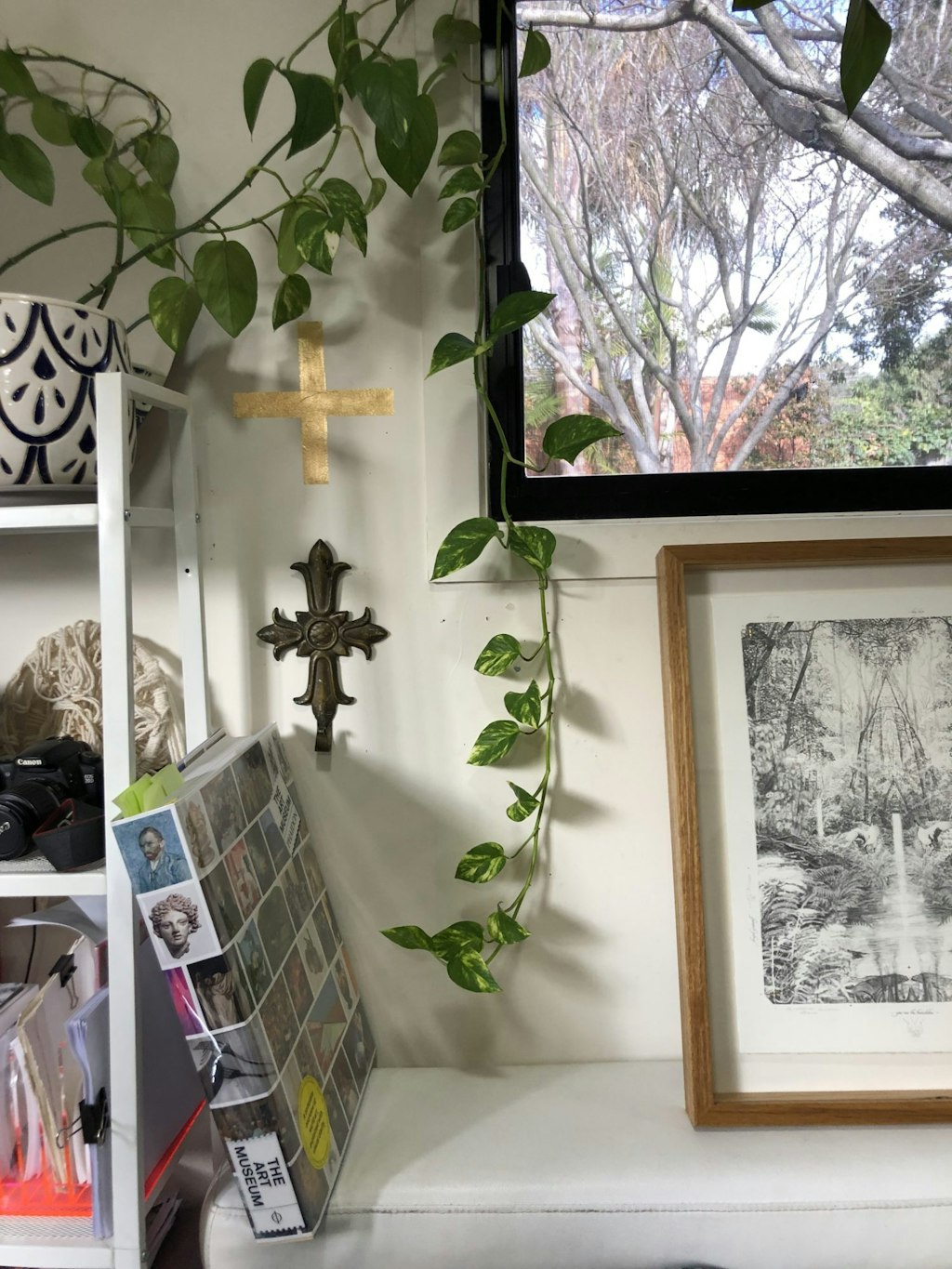 A shelving unit holds objects including a pot plant trailing leaves. Between the unit and a window, two crucifixes hang on a wall. A book and a framed artwork sit on a piece of furniture.