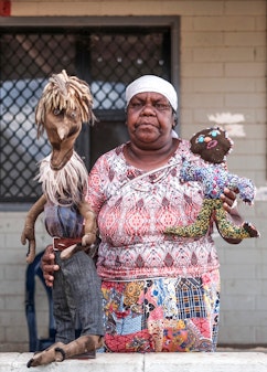 An Aboriginal woman stands holding a soft sculpture of a figure in each hand.