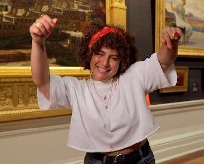 In a gallery space with historic paintings on the wall, a smiling person holds their arms in the air, fingers pointing down.