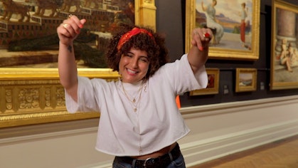 In a gallery space with historic paintings on the wall, a smiling person holds their arms in the air, fingers pointing down.