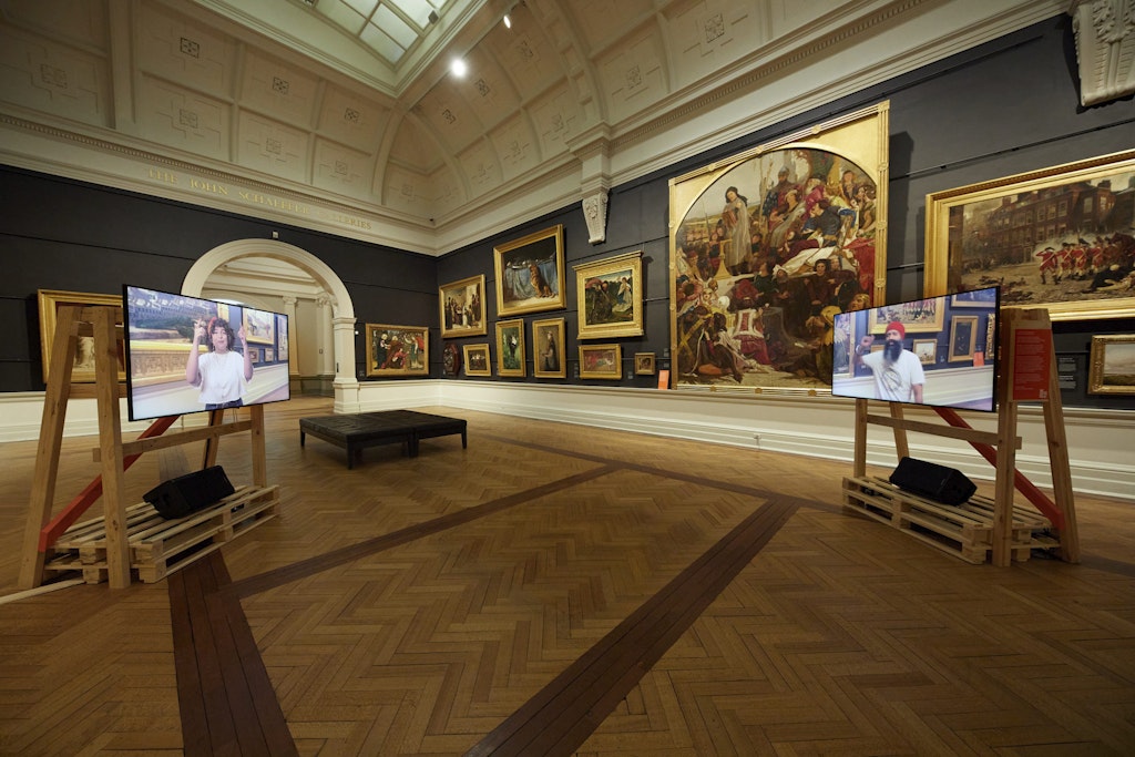 Two video screens on woodern stands sit in an historic gallery space where gold-framed paintings hang on the walls. On each screen is a different person filmed in that gallery.