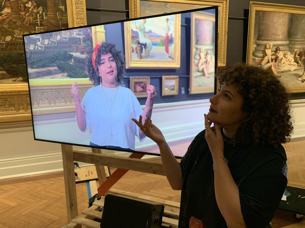 In a gallery space with historic paintings on the wall, a person stands next to a video screen on which they are shown in that same gallery.