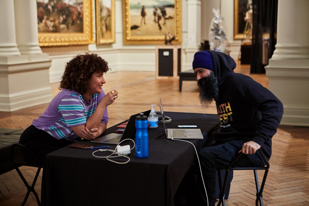 In a gallery space displaying historic artworks, two people sit around a table with laptop computers.