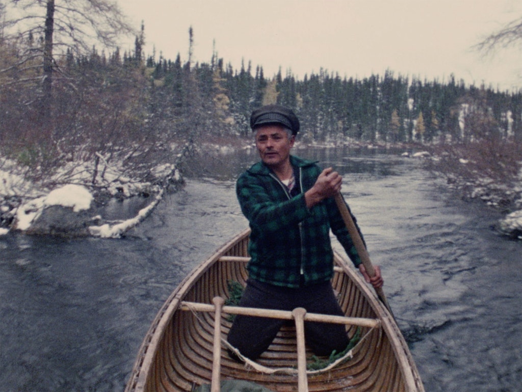 A person kneels in a canoe, paddling it down a river, with snow-covered rocks and leafless trees on the banks.