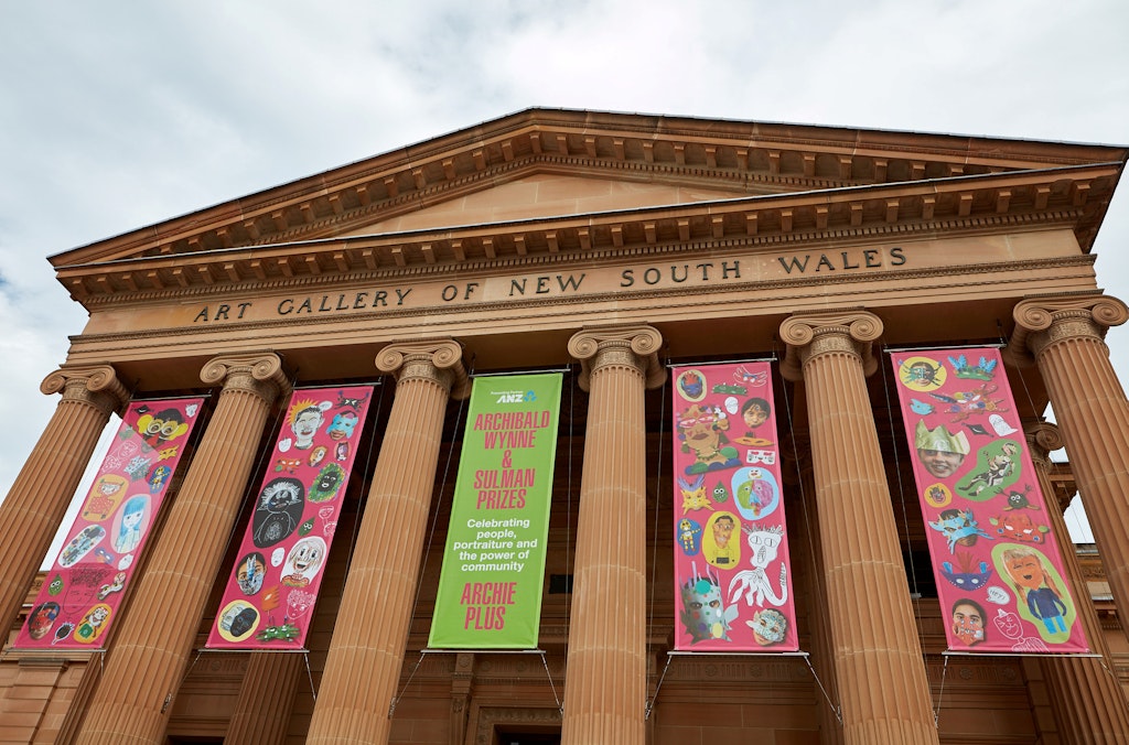 Five banners hang between sandstone columns at the front of the building with Art Gallery of New South Wales on the facade. Four of the banners feature colourful artwork.