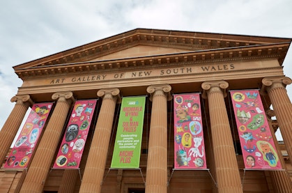 Five banners hang between sandstone columns at the front of the building with Art Gallery of New South Wales on the facade. Four of the banners feature colourful artwork.