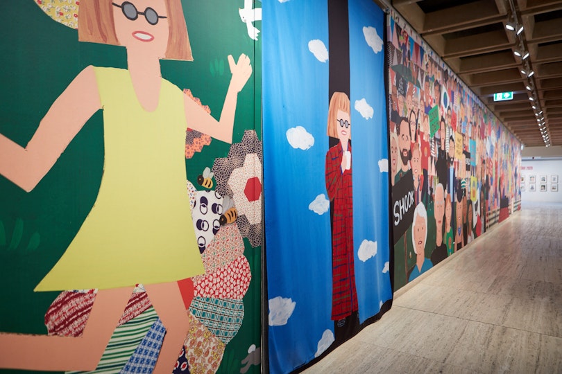 A long mural features a person with bobbed red hair and glasses running in a yellow dress and peaking out from curtains dressed in red. The rest of the wall features many different people.