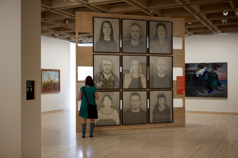 A person looks at nine head-and-torso portraits hung in a grid. Paintings hang on the wall in the background.