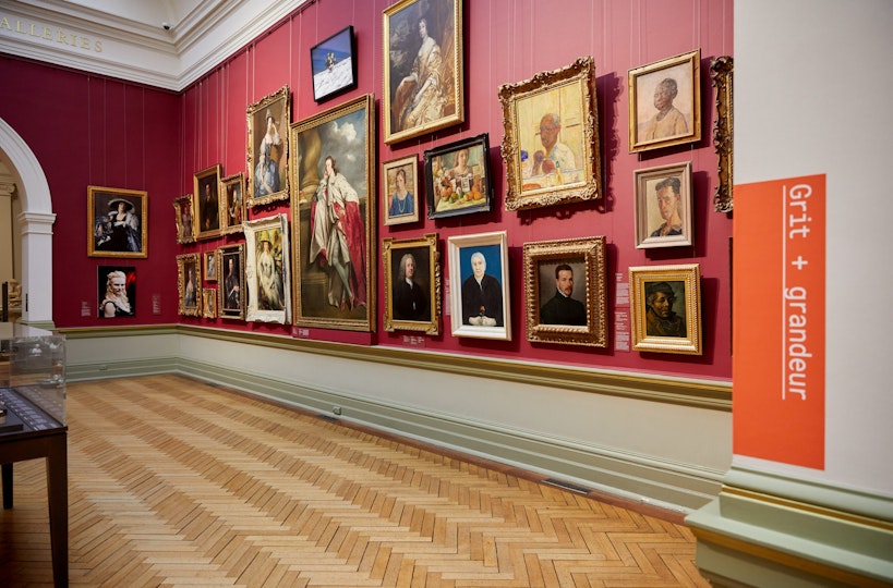 23 framed portrait paintings hang on a red wall next to a sign saying 'Grit + grandeur'.