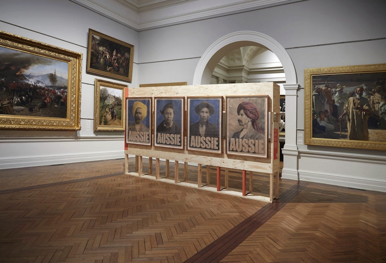 Four head-and-shoulder portraits, each with 'Aussie' in text at the bottom, are hung on a wooden display stand within an historic gallery space displaying framed paintings.