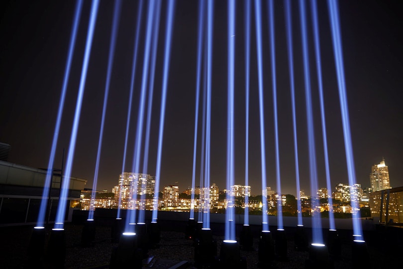 Multiple beams of purple light shine directly up into the night sky. In the background is a cityscape of buildings with lit windows.