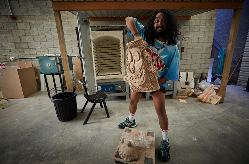 In a room with a few furnishings and scattered materials, a person stands holding a pottery head.