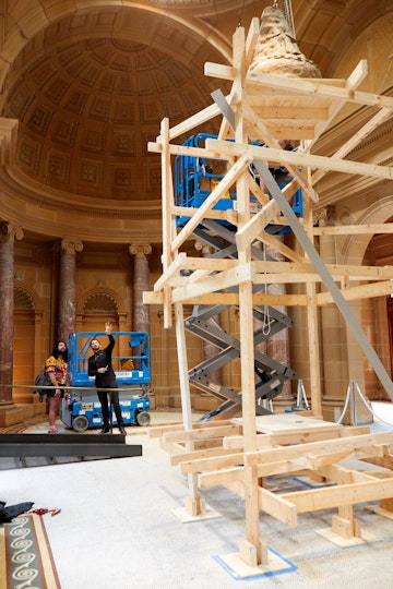 Two people stand in a vaulted room next to wooden scaffolding that holds a ceramic figure.